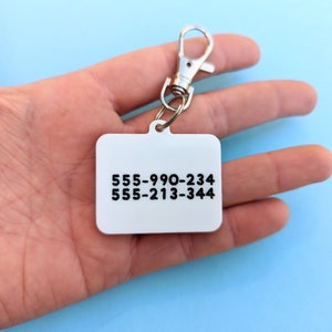 May include: A silver key chain with a white rectangular tag. The tag has two phone numbers printed on it: 555-990-234 and 555-213-344.