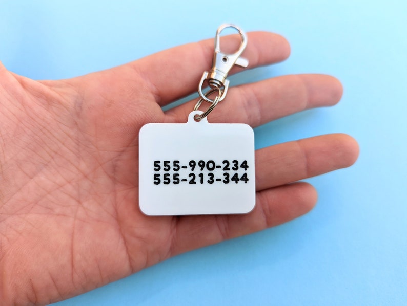 May include: A silver key chain with a white rectangular tag. The tag has two phone numbers printed on it: 555-990-234 and 555-213-344.