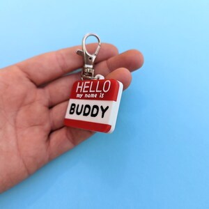 May include: A red and white plastic name tag with a silver key ring. The tag says "HELLO my name is BUDDY".