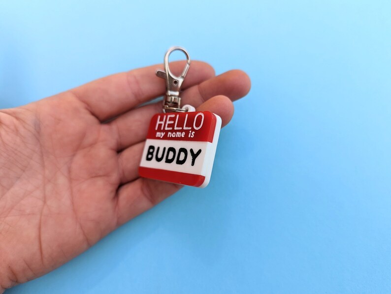 May include: A red and white plastic name tag with a silver key ring. The tag says "HELLO my name is BUDDY".