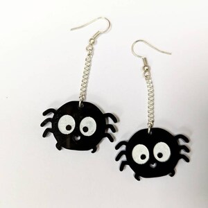 May include: A pair of black spider earrings with white eyes and a smiling mouth. The spider charms hang from silver-colored chain and ear hooks. The earrings are a fun, whimsical accessory.