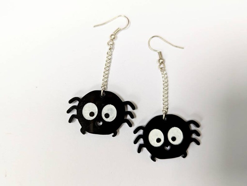 May include: A pair of black spider earrings with white eyes and a smiling mouth. The spider charms hang from silver-colored chain and ear hooks. The earrings are a fun, whimsical accessory.