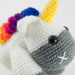 May include: A crocheted unicorn toy with a rainbow mane and a gray face. The unicorn has a yellow horn and black eyes.