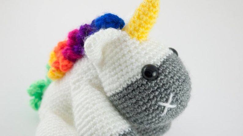 May include: A crocheted unicorn toy with a rainbow mane and a gray face. The unicorn has a yellow horn and black eyes.