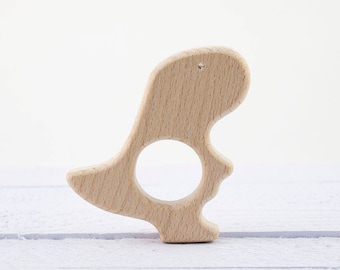 May include: A light-colored wooden dinosaur-shaped baby teether with a circular opening. The teether is made of smooth, unfinished wood and has a small hole at the top. The toy is designed for babies to chew on and is approximately 4 inches tall.