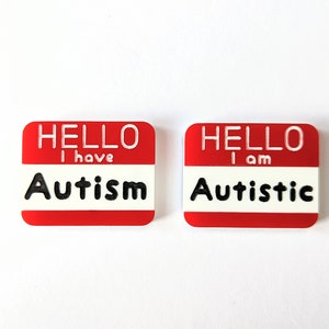 May include: Two red name tags with white lettering. The left tag says "HELLO I have Autism". The right tag says "HELLO I am Autistic".