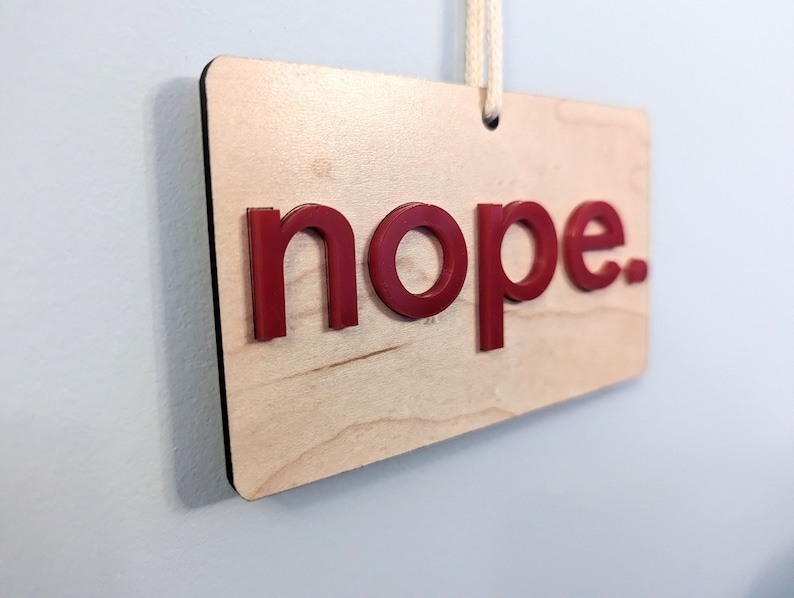 Puede incluir: Un letrero de madera con la palabra "nope" en letras rojas. El letrero est&aacute; colgado de una cuerda blanca.