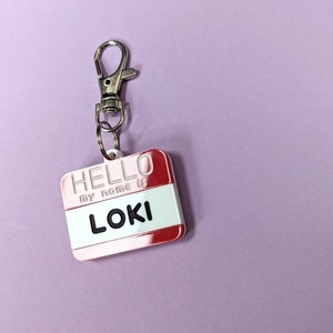 May include: A red and white name tag with a silver keychain. The tag says "HELLO my name is LOKI".
