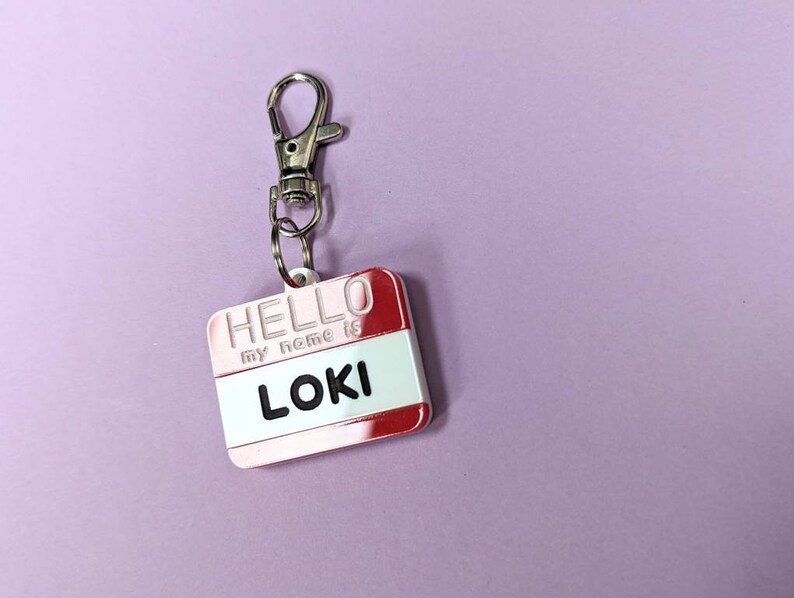 May include: A red and white name tag with a silver keychain. The tag says "HELLO my name is LOKI".