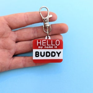 May include: Red and white plastic keychain with a silver ring. The keychain has the text "HELLO my name is BUDDY" on it.
