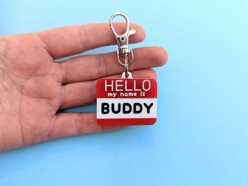 May include: Red and white plastic keychain with a silver ring. The keychain has the text "HELLO my name is BUDDY" on it.
