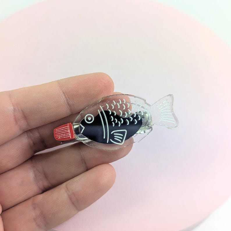 May include: A clear, fish-shaped soy sauce container with a red cap. The container has a black interior with white details, including scales and fins. The design is reminiscent of Japanese cuisine.