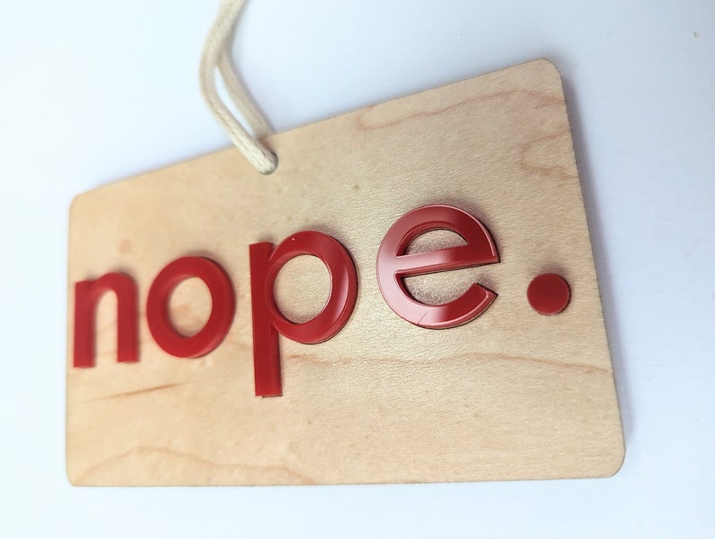 Puede incluir: Un letrero de madera con la palabra "nope" en letras rojas. El letrero tiene una cuerda atada en la parte superior para colgarlo.