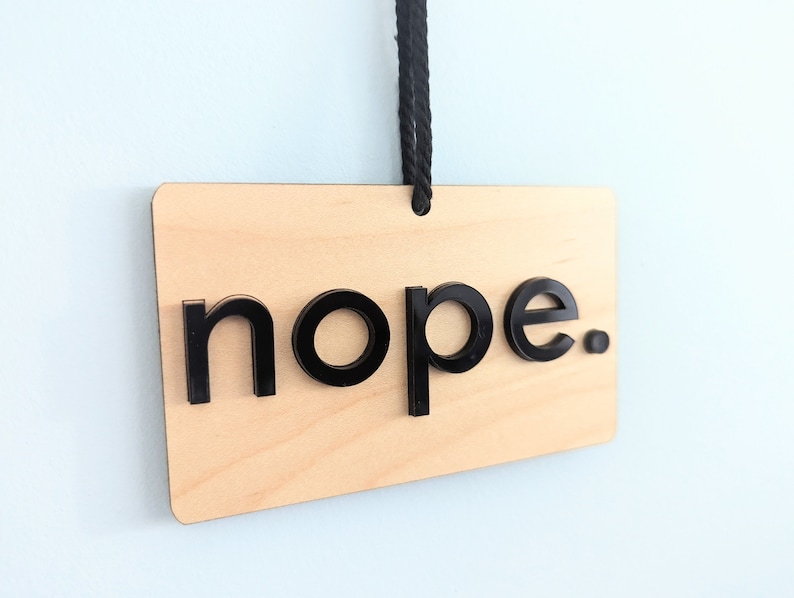 Puede incluir: Un letrero de madera con la palabra "nope" en letras negras. El letrero est&aacute; colgado de una cuerda negra.