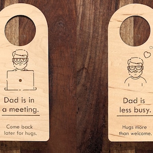 Puede incluir: Dos colgadores de puerta de madera con ilustraciones de una persona con gafas y barba. El primer colgador dice "Papá está en una reunión. Vuelve más tarde para abrazos." El segundo colgador dice "Papá está menos ocupado. Los abrazos son más que bienvenidos."