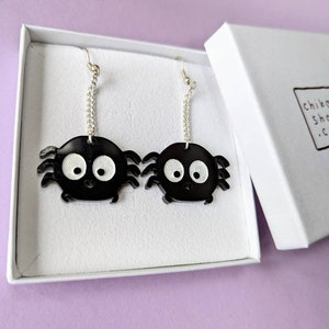 May include: A pair of black spider earrings with white eyes and silver chain attachments are displayed inside a white box. The earrings have a cartoonish design. The box has a logo that reads "chikai shop かい .com".