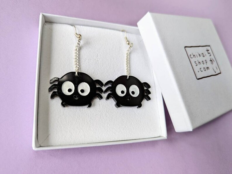 May include: A pair of black spider earrings with white eyes and silver chain attachments are displayed inside a white box. The earrings have a cartoonish design. The box has a logo that reads "chikai shop かい .com".