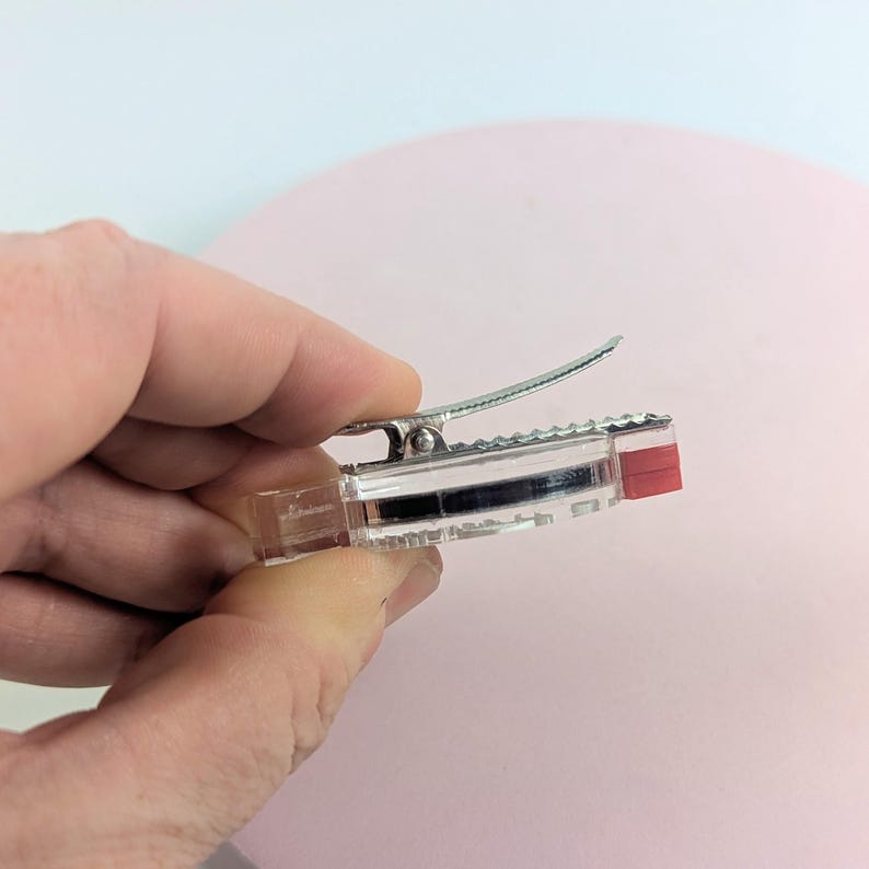 May include: A clear plastic hair clip with a silver metal clip and a red accent on one end. The clip is held in a hand against a pink background. The clip is designed to hold hair in place.