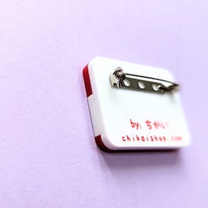 May include: White and red rectangular pin with a silver pin back. The pin has the text "by: ちかい shop" and "chikaishop.com" printed in red.