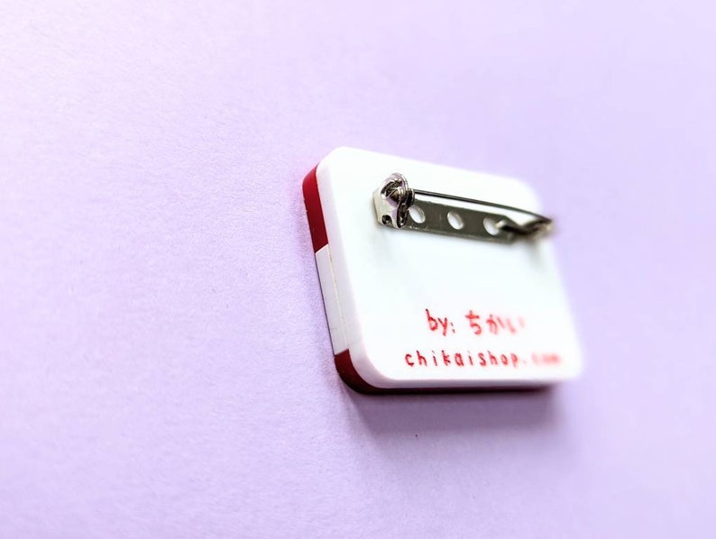 May include: White and red rectangular pin with a silver pin back. The pin has the text "by: ちかい shop" and "chikaishop.com" printed in red.