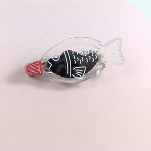 May include: A clear, fish-shaped soy sauce container with a black fish design inside. The container has a red cap and is made of clear plastic. The fish design has white details. The container is on a pink surface.