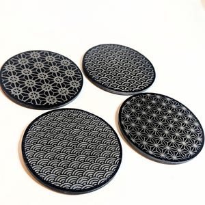 Puede incluir: Cuatro posavasos negros y grises con diferentes patrones geométricos. Los posavasos son redondos y presentan diseños que incluyen una explosión de estrellas, un patrón de olas y un patrón floral geométrico. Los posavasos están sobre una superficie blanca.