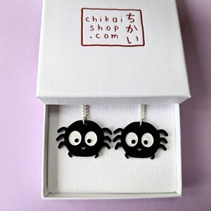 May include: A pair of black spider earrings with white eyes and a smiling mouth, presented in a white box. The earrings hang from silver chains. The box has a red logo with Japanese text.