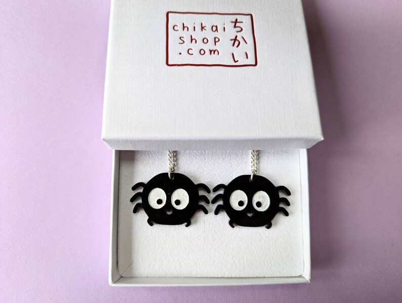 May include: A pair of black spider earrings with white eyes and a smiling mouth, presented in a white box. The earrings hang from silver chains. The box has a red logo with Japanese text.