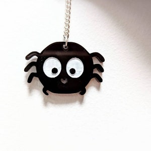 May include: A black spider pendant with white eyes and a smiling mouth hangs from a silver chain. The spider has eight legs and a round body. The pendant is a cute, cartoon-like design.