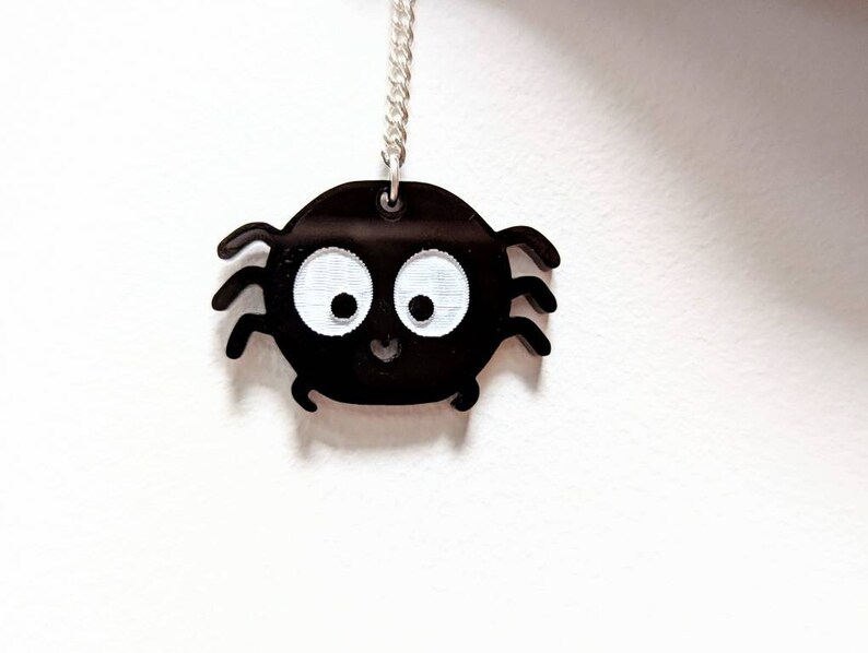 May include: A black spider pendant with white eyes and a smiling mouth hangs from a silver chain. The spider has eight legs and a round body. The pendant is a cute, cartoon-like design.