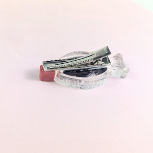 May include: A clear, decorative hair clip with a silver metal clasp. The clip is set on a clear, ridged base with a small red accent. The background is a soft pink.