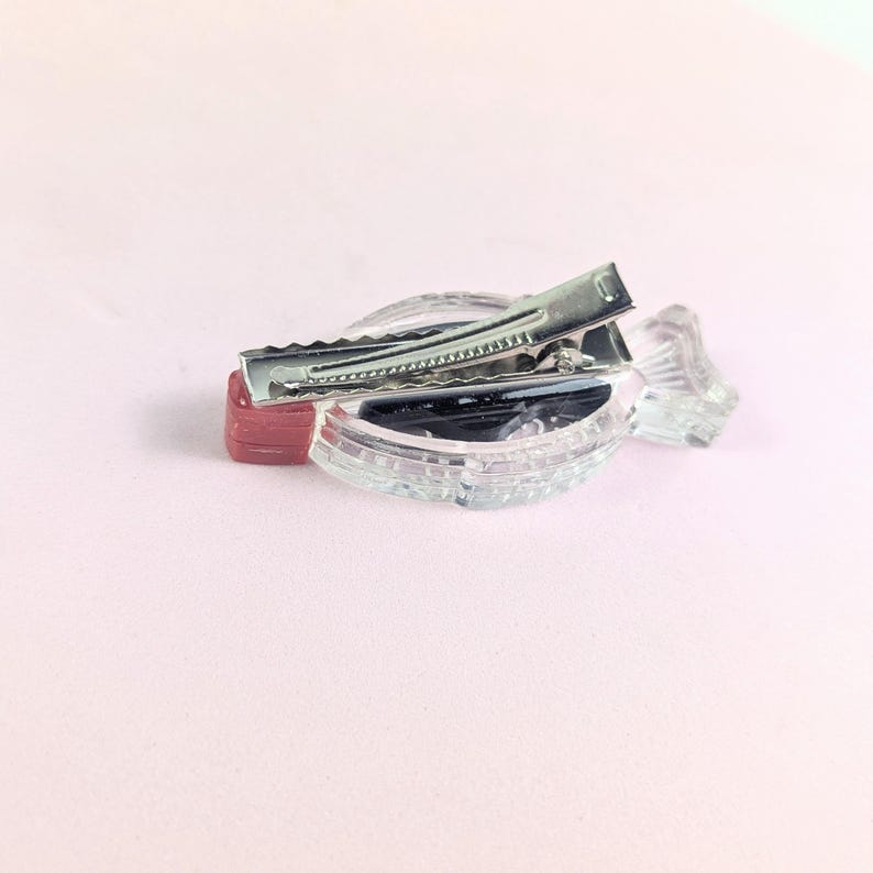 May include: A clear, decorative hair clip with a silver metal clasp. The clip is set on a clear, ridged base with a small red accent. The background is a soft pink.