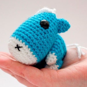 May include: A small crocheted blue and white animal toy with black eyes. The toy is sitting in the palm of a hand.