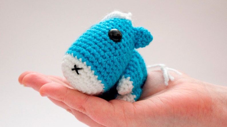 May include: A small crocheted blue and white animal toy with black eyes. The toy is sitting in the palm of a hand.