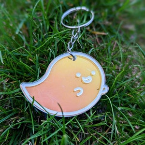 Può includere: Un portachiavi a forma di fantasma giallo e arancione con un viso sorridente. Il portachiavi è in acrilico e ha un anello di metallo argentato.
