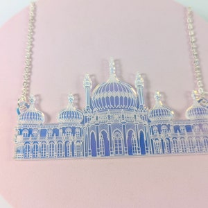 May include: A silver chain necklace with a clear acrylic pendant featuring a detailed outline of a building with multiple domes. The building is a light blue colour with white details.