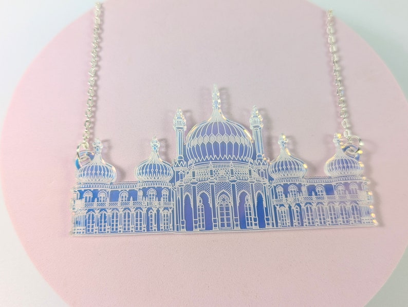 May include: A silver chain necklace with a clear acrylic pendant featuring a detailed outline of a building with multiple domes. The building is a light blue colour with white details.