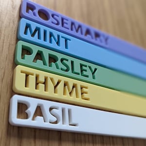 May include: Five colourful plant markers with the names "Rosemary", "Mint", "Parsley", "Thyme", and "Basil" written in white.