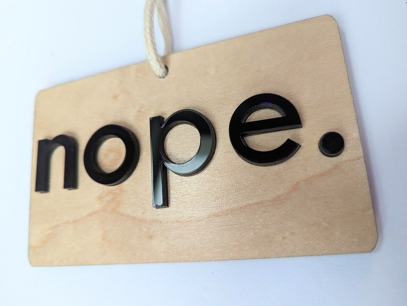 Puede incluir: Un letrero de madera con la palabra "nope" en letras negras en relieve. El letrero tiene un acabado de madera natural y un peque&ntilde;o punto negro despu&eacute;s de la palabra "nope".