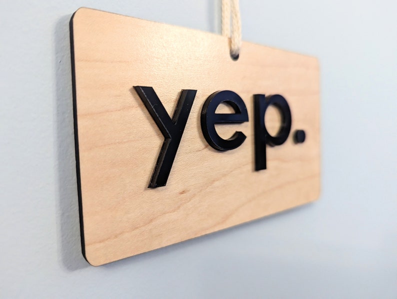 Puede incluir: Un letrero de madera con la palabra "yep." en letras negras en relieve. El letrero tiene una cuerda atada en la parte superior para colgarlo.