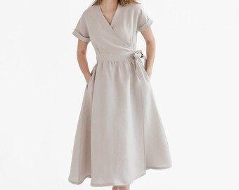 May include: A woman wearing a light beige linen wrap dress with short sleeves and a tie belt. She is smiling and standing with her hands in her pockets.