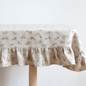 May include: A beige linen tablecloth with a floral print and a ruffled edge. The tablecloth is laid on a wooden table.