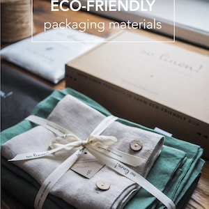 May include: A set of two linen fabric towels, one green and one beige, are folded and tied together with a white ribbon. The towels are packaged in a box with the text "so linen!" on the side. The box is sitting on a wooden surface.