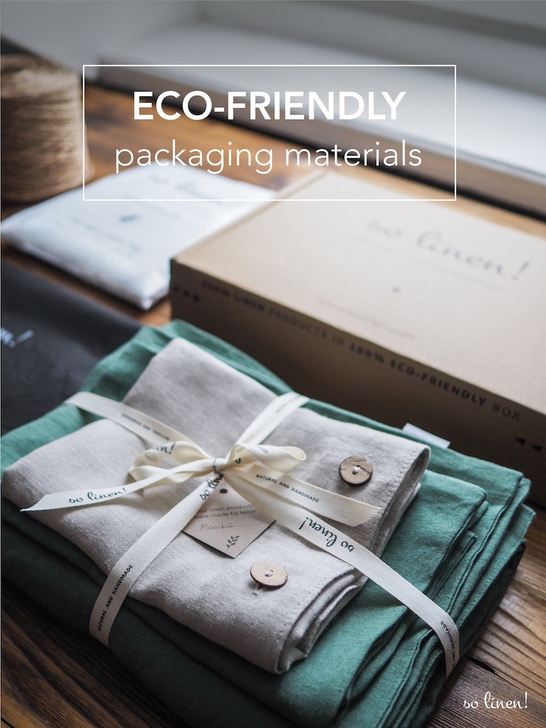 May include: A set of two linen fabric towels, one green and one beige, are folded and tied together with a white ribbon. The towels are packaged in a box with the text "so linen!" on the side. The box is sitting on a wooden surface.