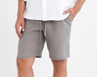 Op de afbeelding: Een persoon draagt een wit overhemd met knopen en een grijze short met een trekkoord. De short komt tot de knie en is gemaakt van een textuurstof. Het overhemd heeft lange mouwen en een relaxte pasvorm.