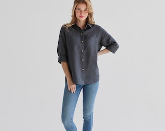May include: A woman wearing a dark gray linen shirt with a button-down collar and long sleeves. She is also wearing blue jeans and is standing on a white background.