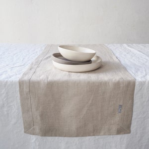 Op de afbeelding: Een beige linnen tafelloper met een subtiele textuur. De loper ligt op een wit linnen tafelkleed. Drie witte keramische kommen zijn in het midden van de loper op elkaar gestapeld.