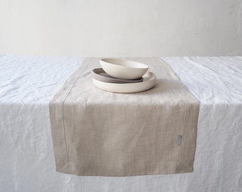 Puede incluir: Un camino de mesa de lino beige con un borde cosido, colocado sobre un mantel blanco. Dos cuencos de cerámica, uno gris y otro blanquecino, están apilados sobre un plato a juego. La escena se desarrolla contra una pared blanca lisa, creando una estética minimalista.