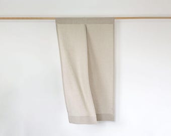 Puede incluir: Una cortina de lino beige cuelga de una barra de madera contra una pared blanca. La cortina está doblada por la mitad, con un borde beige más oscuro en la parte inferior y superior. La tela se drapea suavemente, creando pliegues.