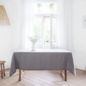 May include: A gray linen tablecloth draped over a wooden table in a bright, airy room with a white wicker pendant light hanging above. The table is set with a white pitcher and flowers.